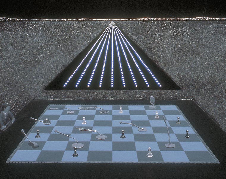 Detail of nightscape with figure seated next to a table on which is a chessboard containing microphones and pawns, focusing on the figure, table, and lights