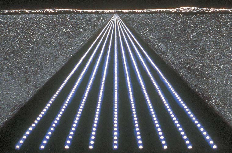 Detail of nightscape with figure seated next to a table on which is a chessboard containing a mixture of microphones and pawns, focusing on the rows of lights that extend to a mountainous horizon